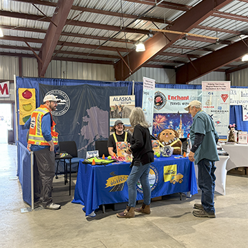 An Alaska DOT&PF promotional booth manned by two staff interacting with two visitors.
