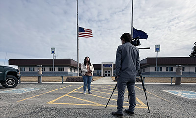 foreground video-camera operator midground information officer holding microphone background flags at half-mast in front of a building