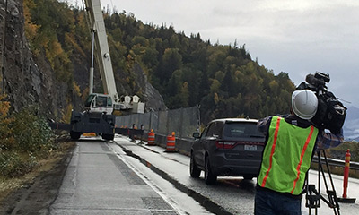 foreground video-camera operator midground parked car background heavy equipment extends to work on a rock wall