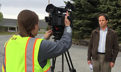 foreground video-camera operator midground parked car background heavy equipment extends to work on a rock wall