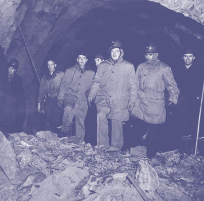View of a group of people stepping over a pile of rubble in the Whittier Tunnel (Anton Anderson Memorial Tunnel) during its construction. The woman on the far left in front of the group is identified as Benzie Ola 'Rusty' Dow. The man at the front and center of the group is General Simon Bolivar Buckner, and to his right is Colonel Benjamin Talley. Nov. 1942