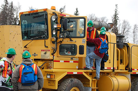 visitors examine a heavy equipment vehicle from its doorway, several other people are watching from the ground