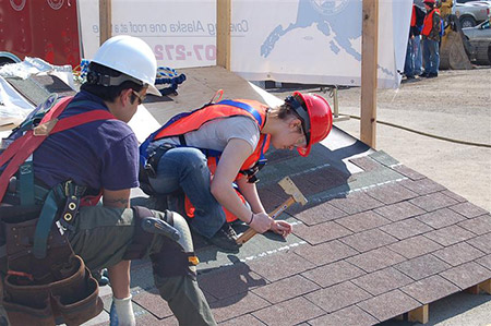 two attendees try their hand at roofing with asphalt shingles, nails, and a hammer at ground level.